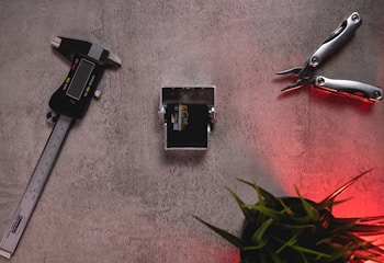 A digital caliper, a small mechanical part, and a multi-tool are arranged on a textured surface. The image also includes some green plant leaves at the bottom right corner with a red light casting a soft glow.