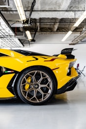 A close-up view of a yellow sports car's rear section inside a garage. The focus is on the rear wheel with intricate alloy design and a visible brake caliper. Overhead, industrial lights hang from the ceiling, adding to the modern aesthetic.