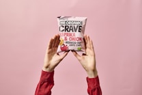 A person wearing a long-sleeved red shirt holds up a bag of organic paprika and onion lentil chips against a soft pink background. The package prominently displays the brand name and flavor.