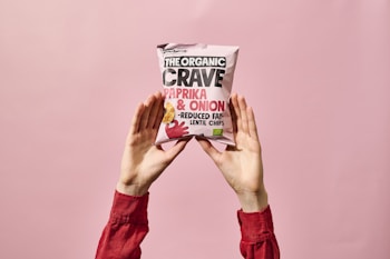 A person wearing a long-sleeved red shirt holds up a bag of organic paprika and onion lentil chips against a soft pink background. The package prominently displays the brand name and flavor.