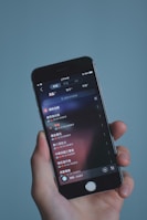 A hand holding a smartphone displaying a music app interface with text in Chinese. The screen shows a list of songs with track durations and additional details. The background is a soft, solid color.
