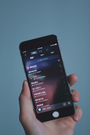 A hand holding a smartphone displaying a music app interface with text in Chinese. The screen shows a list of songs with track durations and additional details. The background is a soft, solid color.