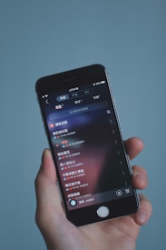 A hand holding a smartphone displaying a music app interface with text in Chinese. The screen shows a list of songs with track durations and additional details. The background is a soft, solid color.
