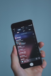 A hand holding a smartphone displaying a music app interface with text in Chinese. The screen shows a list of songs with track durations and additional details. The background is a soft, solid color.