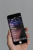 A person holds a smartphone displaying a music application interface, featuring a list of songs in a language that appears to be Chinese. The phone screen shows details such as song titles, artist information, and playback controls. The background is a neutral gray color.