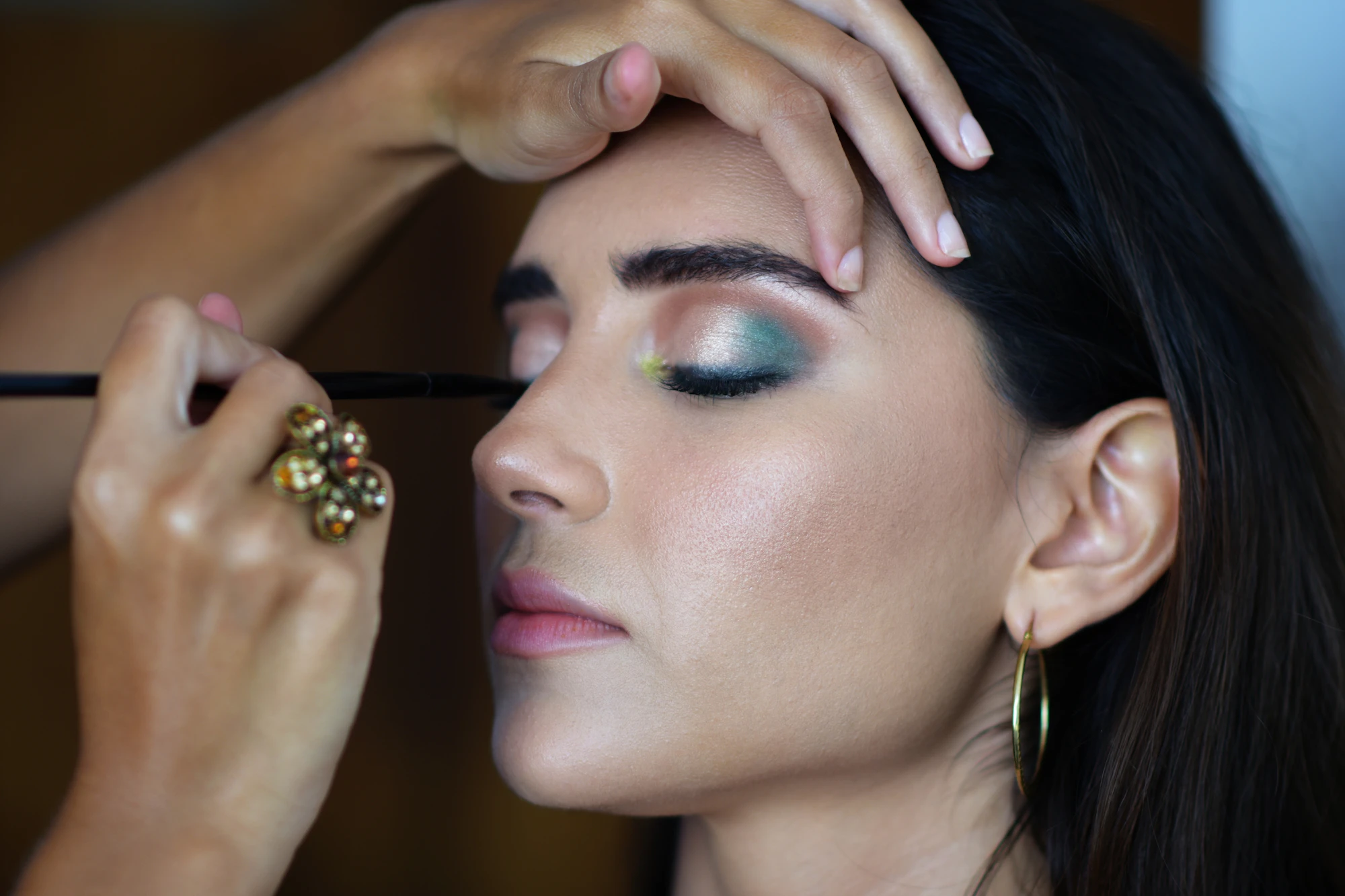 Maquilladora aplicando sombra verde en el párpado de una modelo de piel morena con aretes dorados