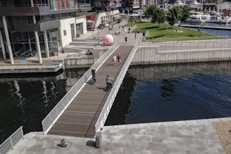 A modern urban area featuring a wooden pedestrian bridge crossing over a body of water. People are walking and cycling on the bridge, with green spaces and trees visible in the background. The architecture is contemporary with glass and concrete buildings along the walkway, and several moored boats are seen on the right side.