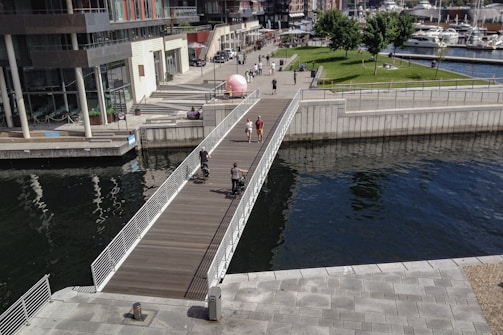 A modern urban area featuring a wooden pedestrian bridge crossing over a body of water. People are walking and cycling on the bridge, with green spaces and trees visible in the background. The architecture is contemporary with glass and concrete buildings along the walkway, and several moored boats are seen on the right side.