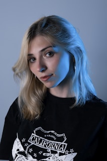 A person with blonde hair and a subtle expression is wearing a black t-shirt with a graphic design featuring the word California. The lighting casts a blue hue on their face and hair, giving a dramatic effect against a plain background.