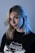 A person with blonde hair and a subtle expression is wearing a black t-shirt with a graphic design featuring the word California. The lighting casts a blue hue on their face and hair, giving a dramatic effect against a plain background.