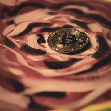 A gold Bitcoin coin is placed on a surface with a swirling pattern of rose and black colors. The intricate design of the pattern adds an artistic flair to the image, contrasting with the smooth, reflective texture of the coin.