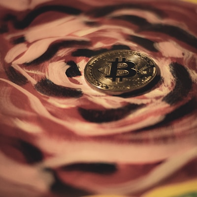 A gold Bitcoin coin is placed on a surface with a swirling pattern of rose and black colors. The intricate design of the pattern adds an artistic flair to the image, contrasting with the smooth, reflective texture of the coin.