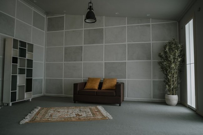 A minimalist living room with a gray geometric wall design and a modern aesthetic. The room includes a brown sofa with two mustard-yellow pillows, a patterned rug, a tall potted plant, and an open shelf with various compartments. Natural light enters from a large window on the right, and a single overhead lamp hangs from the ceiling.