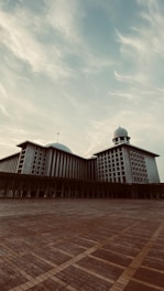 A large mosque with a prominent dome and minaret, showcasing modern architectural style. The foreground features a wide expanse of tiled ground. The sky above is partly cloudy, contributing to a serene atmosphere.