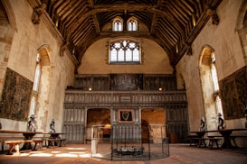 An ornate historical hall with high vaulted ceilings and wooden beams. The walls are adorned with tapestries and suits of armor. Large stained glass windows provide natural light, casting an amber glow on the stone floor. The setting includes long wooden tables positioned along the walls and what appears to be a fireplace surrounded by a protective screen.