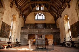 An ornate historical hall with high vaulted ceilings and wooden beams. The walls are adorned with tapestries and suits of armor. Large stained glass windows provide natural light, casting an amber glow on the stone floor. The setting includes long wooden tables positioned along the walls and what appears to be a fireplace surrounded by a protective screen.