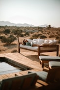 A person relaxes on a cushioned outdoor sofa, surrounded by a serene desert landscape. The setting includes wooden decking and a small pool, with distant mountains under a clear sky.