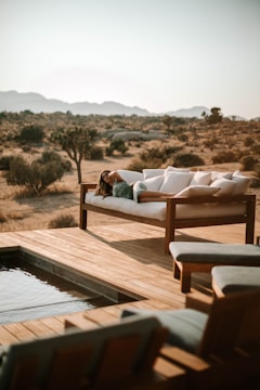 A person relaxes on a cushioned outdoor sofa, surrounded by a serene desert landscape. The setting includes wooden decking and a small pool, with distant mountains under a clear sky.