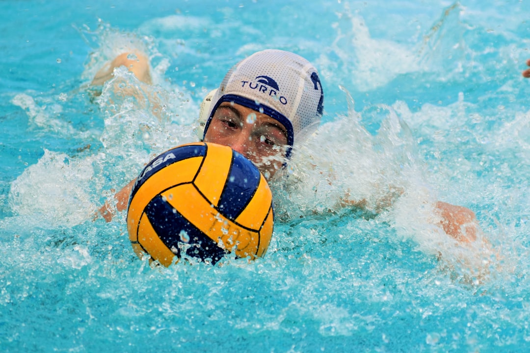 A boy in a white cap, playing a water polo game, swims towards the goals with the ball in front of him. 