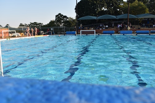 An outdoor swimming pool with clear blue water, bordered by trees and a fence. Several people are gathered on the left side of the pool, while others are seated in a shaded, tiered seating area on the right.