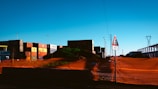 A construction or industrial site with shipping containers stacked in the background. An orange safety netting surrounds a mound of dirt or debris. There is a triangular road sign indicating a cautionary distance of 100 meters. Power lines and a pylon are visible in the distance.