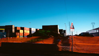 A construction or industrial site with shipping containers stacked in the background. An orange safety netting surrounds a mound of dirt or debris. There is a triangular road sign indicating a cautionary distance of 100 meters. Power lines and a pylon are visible in the distance.