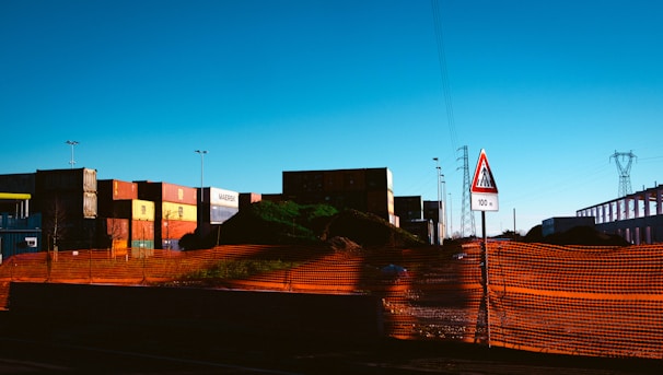 A construction or industrial site with shipping containers stacked in the background. An orange safety netting surrounds a mound of dirt or debris. There is a triangular road sign indicating a cautionary distance of 100 meters. Power lines and a pylon are visible in the distance.
