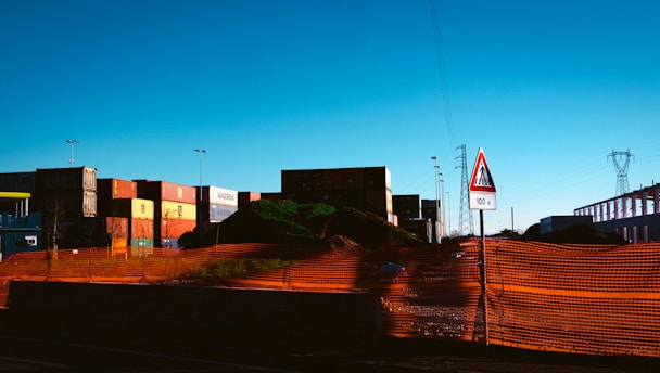 A construction or industrial site with shipping containers stacked in the background. An orange safety netting surrounds a mound of dirt or debris. There is a triangular road sign indicating a cautionary distance of 100 meters. Power lines and a pylon are visible in the distance.