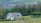 A partially constructed house stands amidst lush greenery, with scaffolding on the front and surrounded by trees. In the background, a residential area with numerous houses stretches across the horizon, nestled among rolling hills and fields.