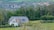 A partially constructed house stands amidst lush greenery, with scaffolding on the front and surrounded by trees. In the background, a residential area with numerous houses stretches across the horizon, nestled among rolling hills and fields.