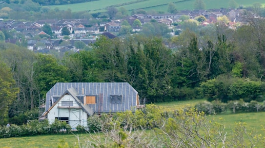 A partially constructed house stands amidst lush greenery, with scaffolding on the front and surrounded by trees. In the background, a residential area with numerous houses stretches across the horizon, nestled among rolling hills and fields.