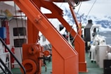 The image displays a close-up view of an industrial or marine setting. Prominent orange metal structures dominate the scene, possibly part of a crane or equipment on a ship deck. The background shows an icy, snow-covered landscape, indicating a cold, possibly polar environment.