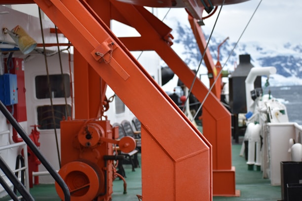 The image displays a close-up view of an industrial or marine setting. Prominent orange metal structures dominate the scene, possibly part of a crane or equipment on a ship deck. The background shows an icy, snow-covered landscape, indicating a cold, possibly polar environment.