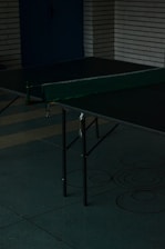 A dimly lit room with a table tennis setup. The table is green with a net across the middle. The flooring is covered with greenish tiles and walls made of white bricks. Circular patterns are present on the floor near the table.