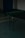 A dimly lit room with a table tennis setup. The table is green with a net across the middle. The flooring is covered with greenish tiles and walls made of white bricks. Circular patterns are present on the floor near the table.