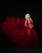 A pregnant woman with long, wavy silver hair is wearing a flowing, voluminous red gown. The fabric of the gown is sheer and creates a dramatic, billowing effect against the dark background. The woman's hands are gently resting on her baby bump, and her expression is serene and contemplative.