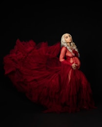A pregnant woman with long, wavy silver hair is wearing a flowing, voluminous red gown. The fabric of the gown is sheer and creates a dramatic, billowing effect against the dark background. The woman's hands are gently resting on her baby bump, and her expression is serene and contemplative.