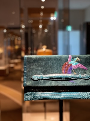 A luxurious handbag with a crocodile texture and a vibrant embroidered bird design, set against a soft, suede background. The bag is prominently displayed in a well-lit room with a blurred background, adding an elegant and high-end feel.