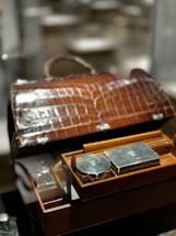 A luxurious brown leather case with a crocodile pattern is open, revealing a neatly organized selection of silver grooming items. The items include a round and a rectangular silver container, embedded within the box's compartments, reflecting light due to their polished surfaces.