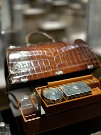 A luxurious brown leather case with a crocodile pattern is open, revealing a neatly organized selection of silver grooming items. The items include a round and a rectangular silver container, embedded within the box's compartments, reflecting light due to their polished surfaces.
