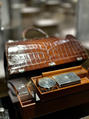 A luxurious brown leather case with a crocodile pattern is open, revealing a neatly organized selection of silver grooming items. The items include a round and a rectangular silver container, embedded within the box's compartments, reflecting light due to their polished surfaces.