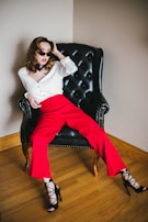 A person is seated on a black leather armchair wearing bold red pants, a white top, and black high-heeled sandals. The person is posing confidently with sunglasses, and their hair styled in loose waves. The room has wooden flooring and a light-colored wall.