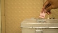 A hand is placing a folded Indonesian banknote into a silver metallic box with a slot on top. The background features a textured golden surface.
