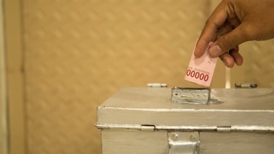 A hand is placing a folded Indonesian banknote into a silver metallic box with a slot on top. The background features a textured golden surface.