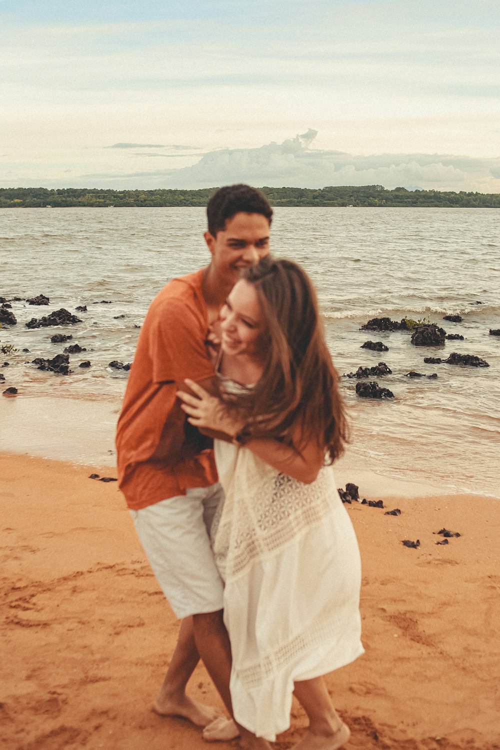 man in orange long sleeve shirt kissing woman in white dress on beach during daytime