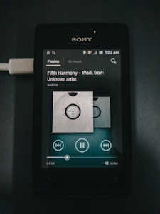A Sony smartphone displaying a music player interface with the song 'Fifth Harmony - Work from Unknown artist'. The screen shows playback controls, album art, and a progress bar at 01:04 of 03:40. The phone is connected to a charger visible on the left side.