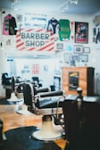A classic barbershop interior with vintage barber chairs and a traditional red and white striped barber shop sign. The walls are adorned with memorabilia including sports jerseys, posters, and framed photos. The wooden floor and warm lighting create a nostalgic atmosphere.