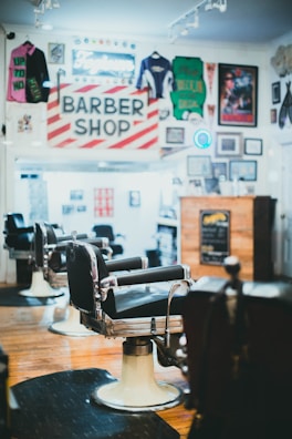 A classic barbershop interior with vintage barber chairs and a traditional red and white striped barber shop sign. The walls are adorned with memorabilia including sports jerseys, posters, and framed photos. The wooden floor and warm lighting create a nostalgic atmosphere.
