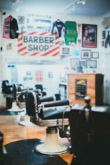 A classic barbershop interior with vintage barber chairs and a traditional red and white striped barber shop sign. The walls are adorned with memorabilia including sports jerseys, posters, and framed photos. The wooden floor and warm lighting create a nostalgic atmosphere.