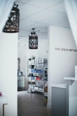 Interior space featuring shelves stocked with various products, likely skincare or cosmetics. The walls are light-colored and the floor is tiled. Large, ornate light fixtures hang from the ceiling. A sign on the wall reads 'The Gold Method'.